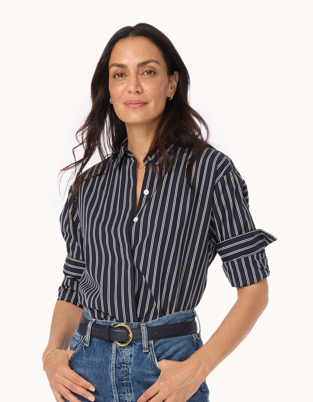 Woman wearing a navy and white striped button down shirt with rolled-up sleeves and blue jeans with a black belt on a white background