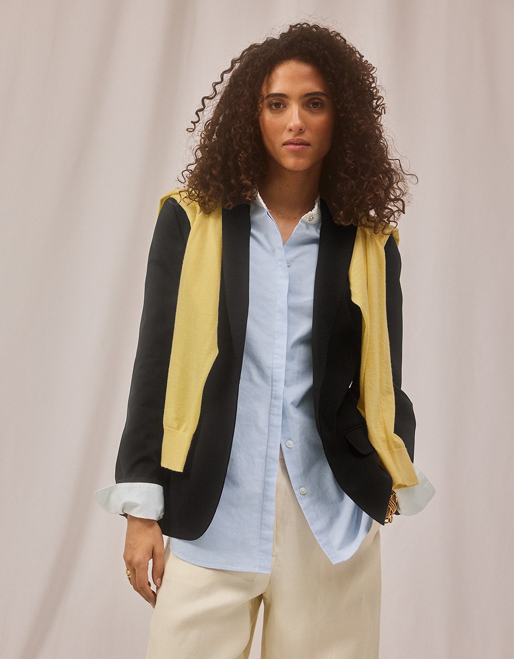 Woman wearing a layered black blazer and draped yellow sweater over a light blue shirt with beige pants on a neutral background