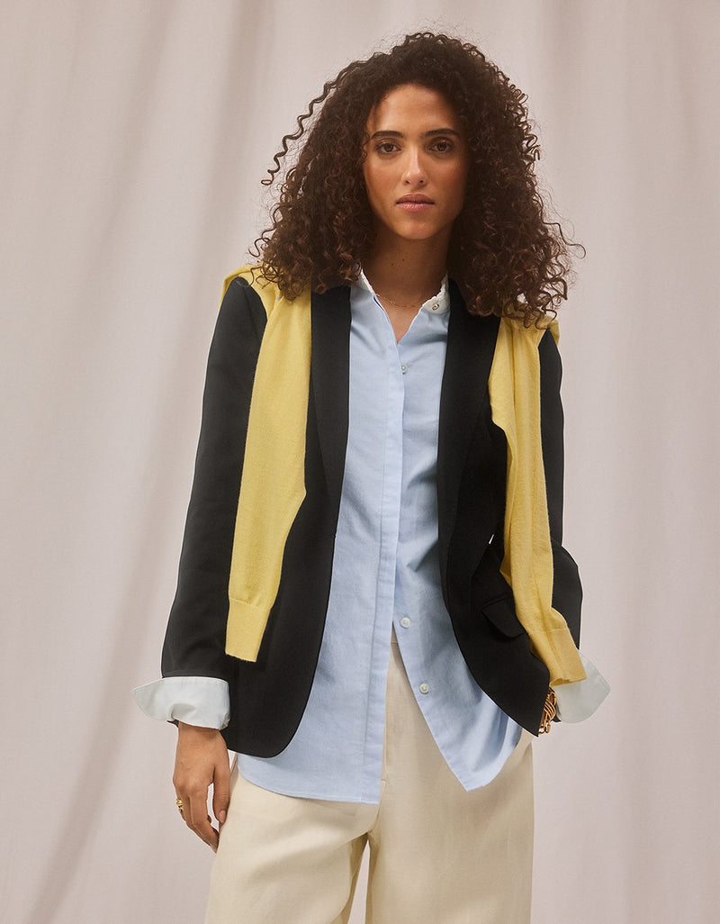 Woman wearing a layered black blazer and draped yellow sweater over a light blue shirt with beige pants on a neutral background