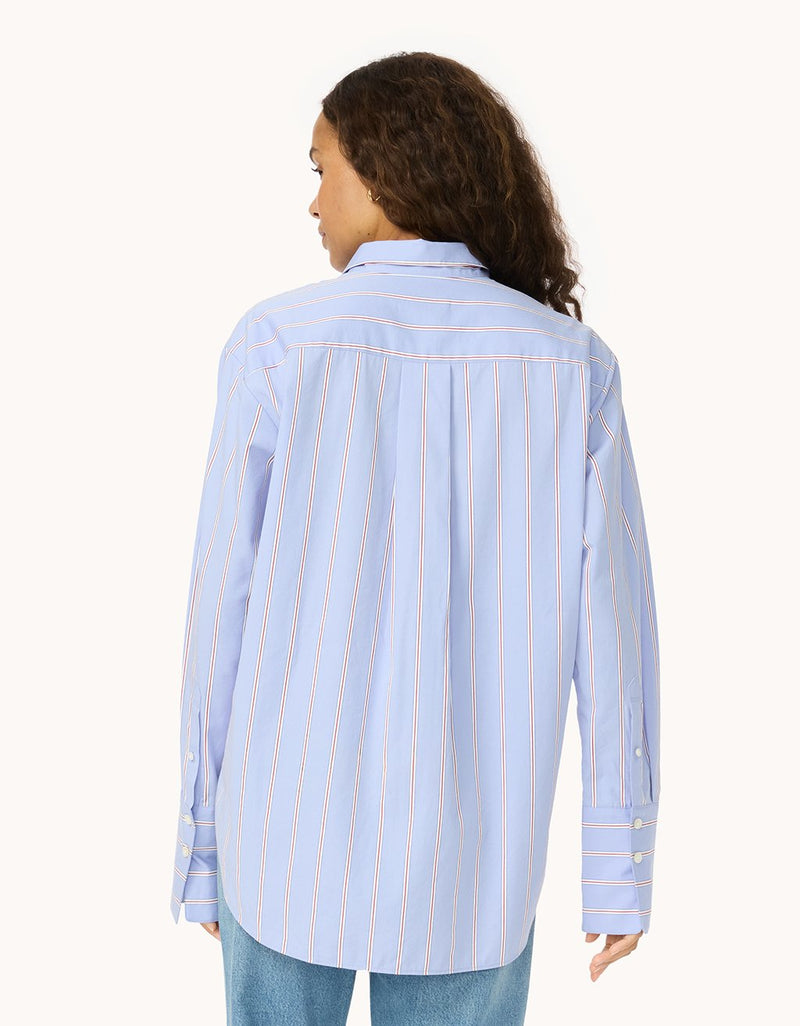 Back view of a woman wearing a light blue  shirt with red and white stripes and blue jeans on a light gray background