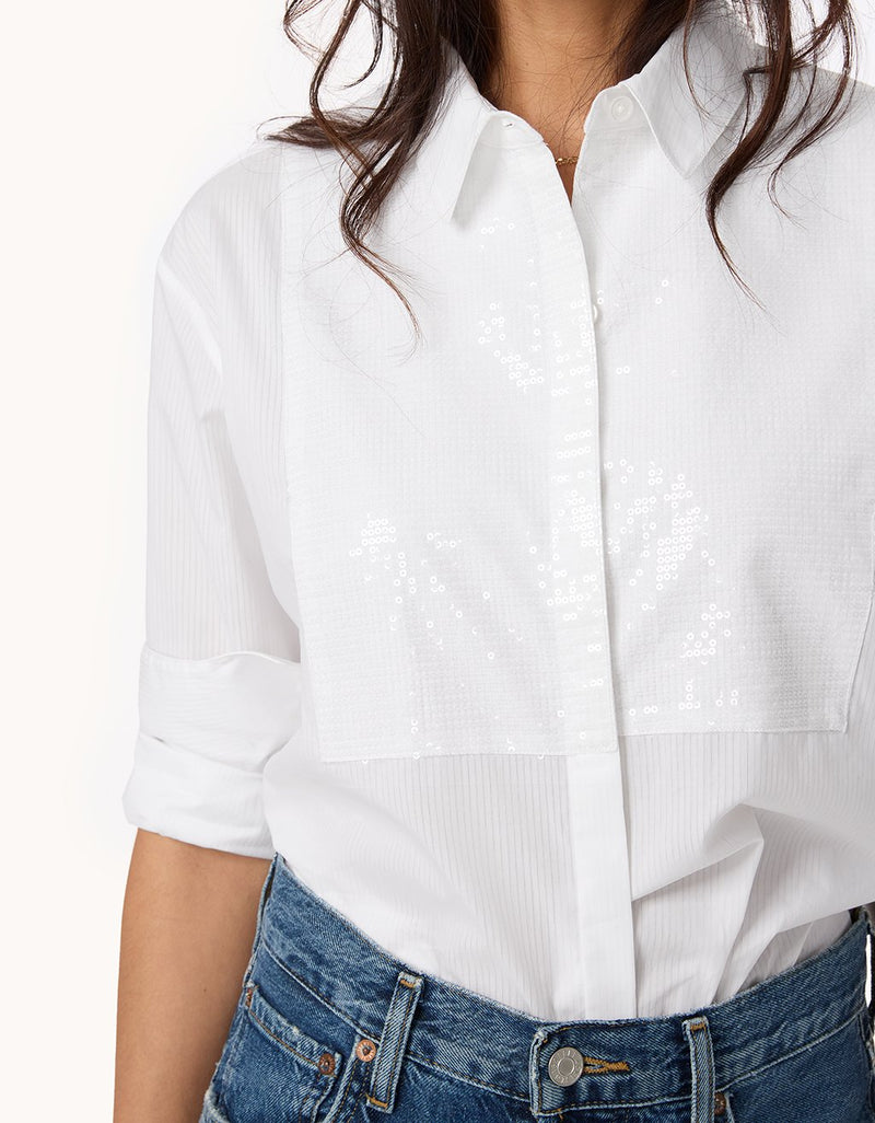 Close-up of a white button down shirt with sequin bib detail and blue jeans worn by a person with a white background
