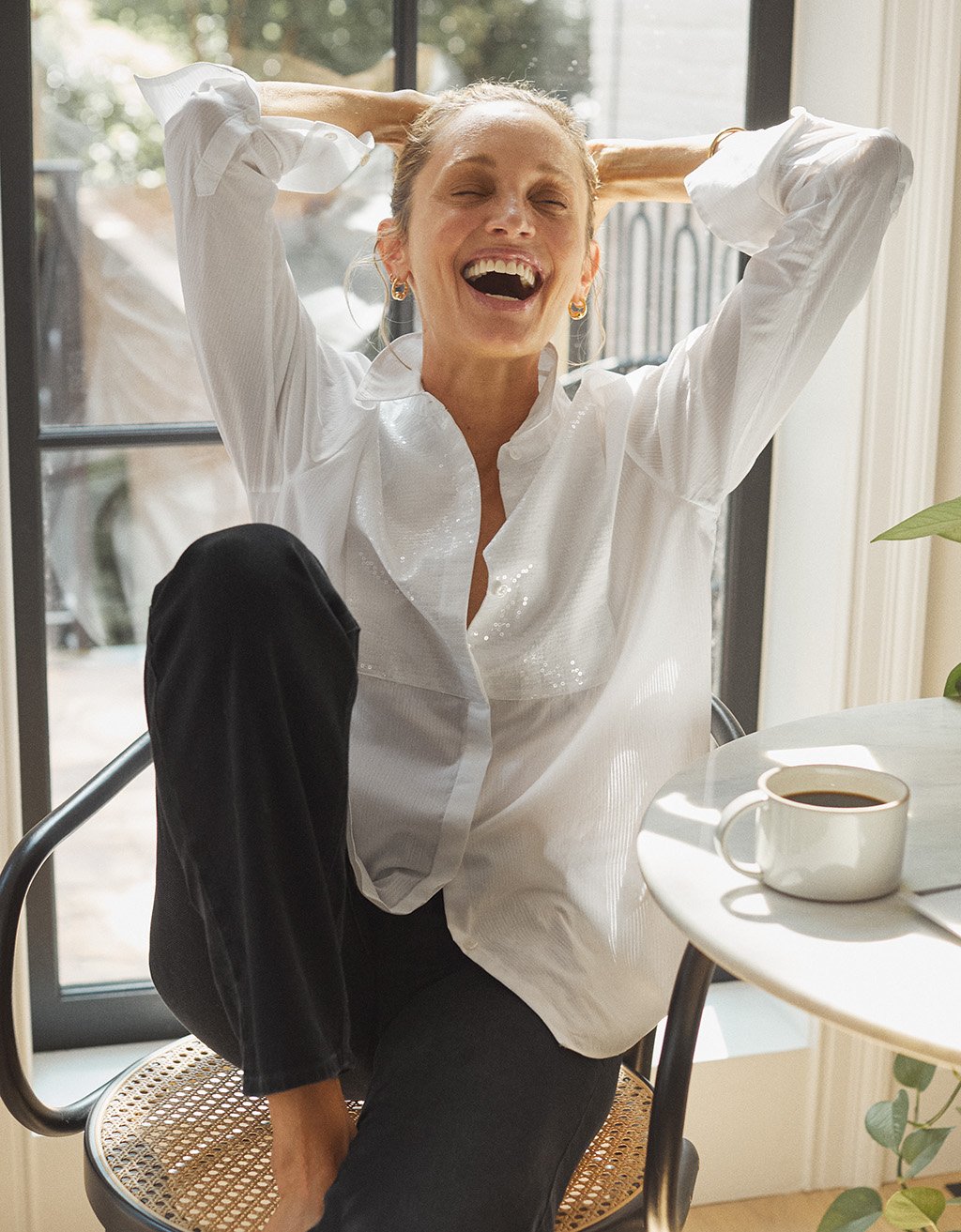 Woman wearing a white button down shirt with sequin bib detail and black pants, sitting on a chair with arms raised, laughing, in a room with a table and mug.