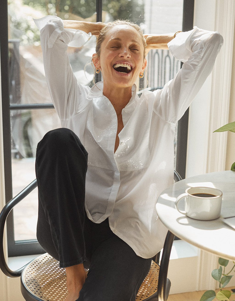 Woman wearing a white button down shirt with sequin bib detail and black pants, sitting on a chair with arms raised, laughing, in a room with a table and mug.