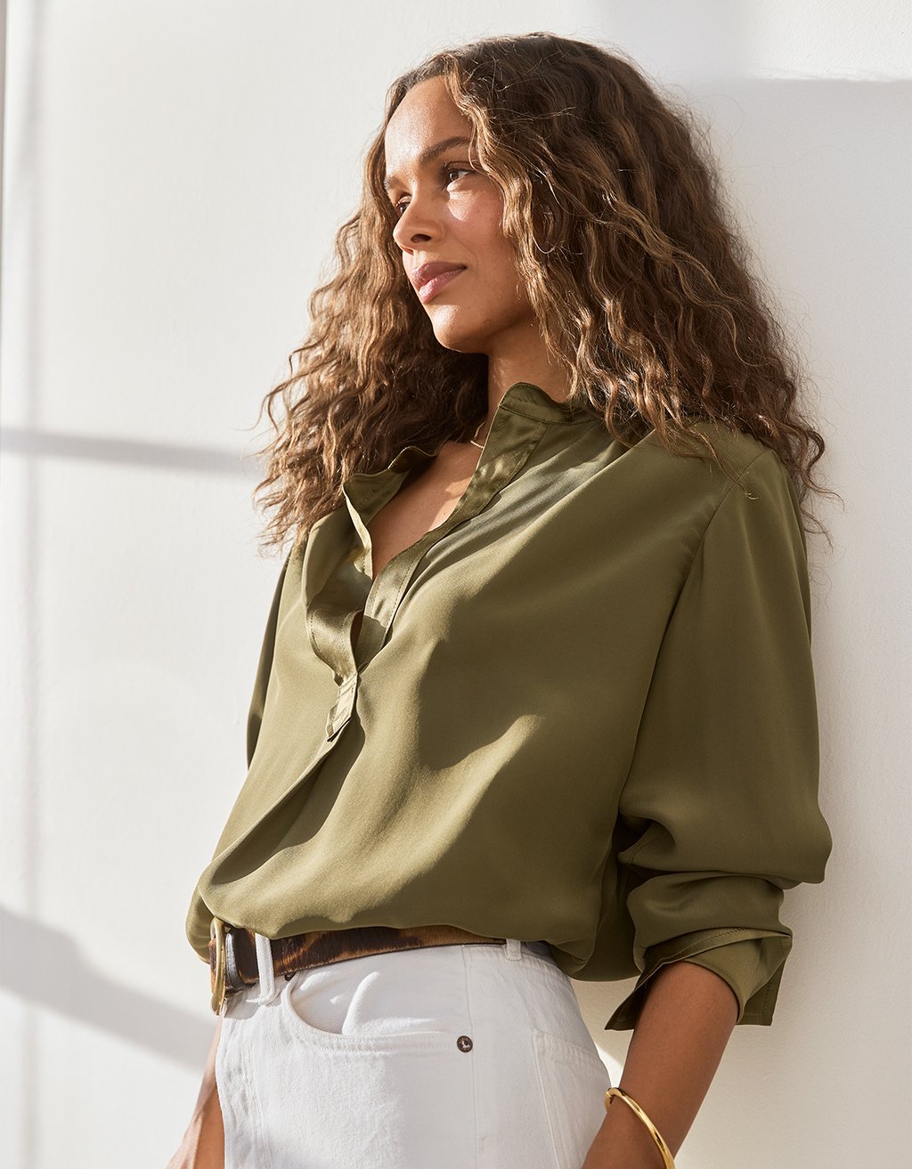 Woman wearing an olive green silk blouse and white jeans against a white wall.