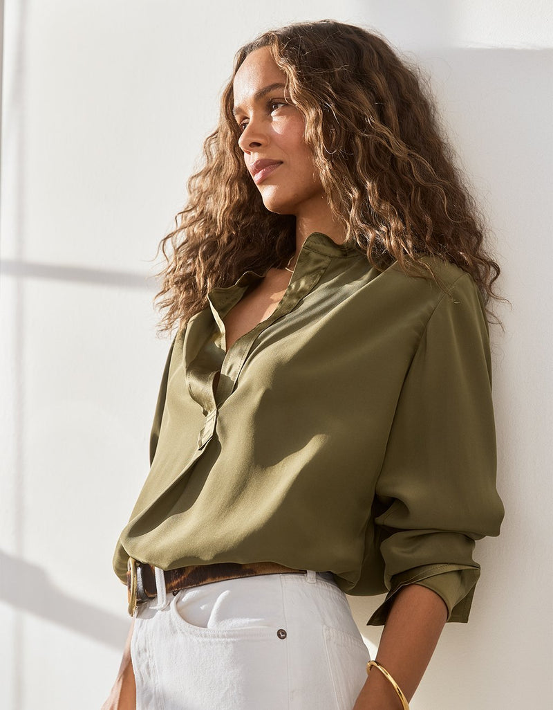 Woman wearing an olive green silk blouse and white jeans against a white wall.