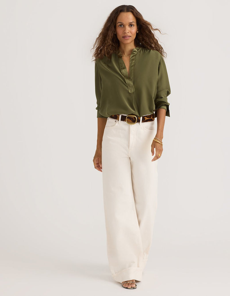 Woman wearing an olive green silk blouse and white pants on a plain background