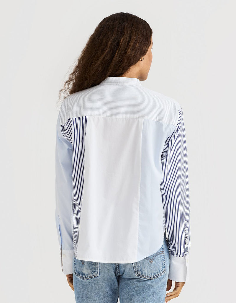 Back view of a woman wearing a striped colorblock tuxedo button down shirt and blue jeans on a plain background