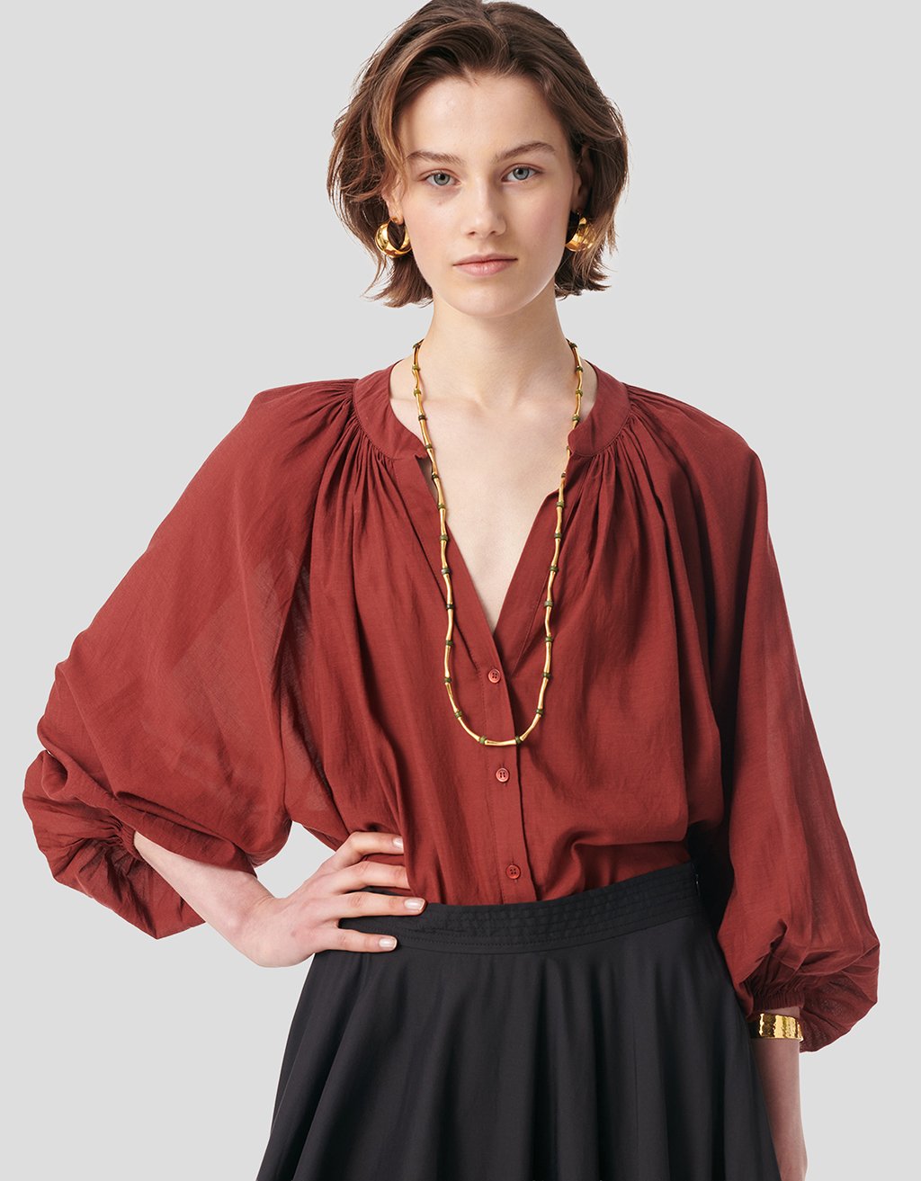 Person wearing a V-neck rust-colored blouse with button down front or balloon sleeves and white pants on a plain background