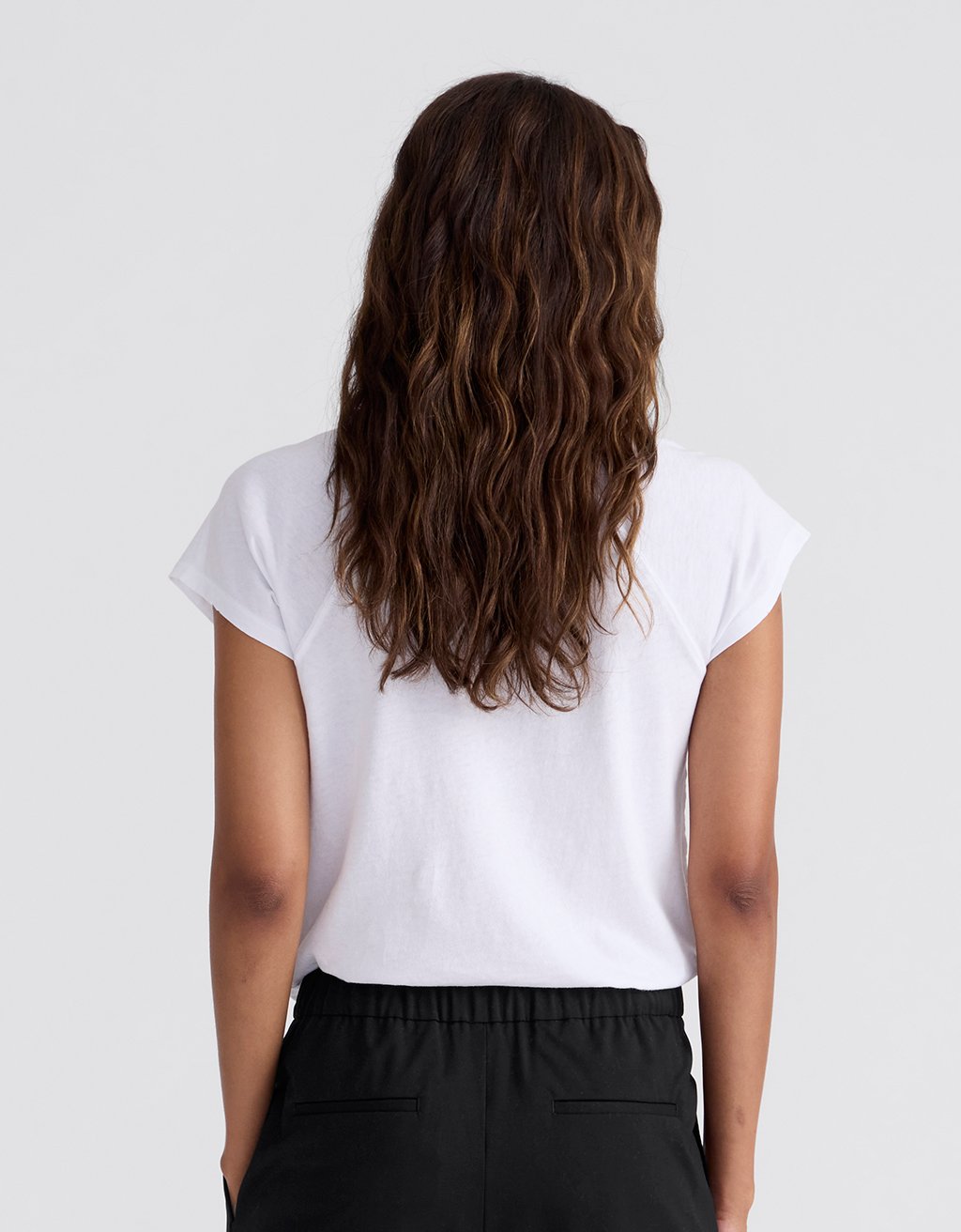Back view of a woman wearing a white raglan t-shirt and gray pants on a plain background