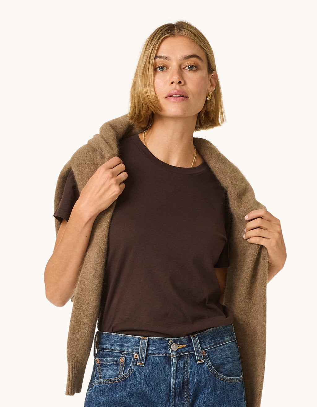 Woman wearing a brown t-shirt, blue jeans, and a brown cardigan draped over her shoulders on a light gray background