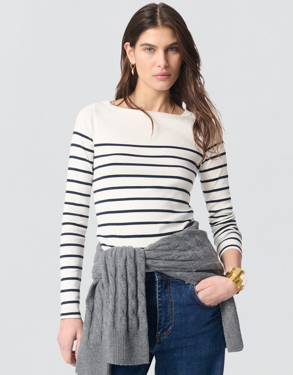 Woman wearing a striped sweater, gray cardigan tied around the waist, and blue jeans on a light gray background
