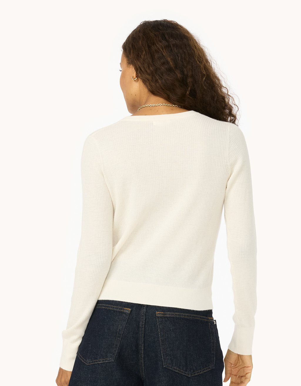 Back view of a woman wearing an ivory ribbed cashmere sweater and dark jeans on a white background