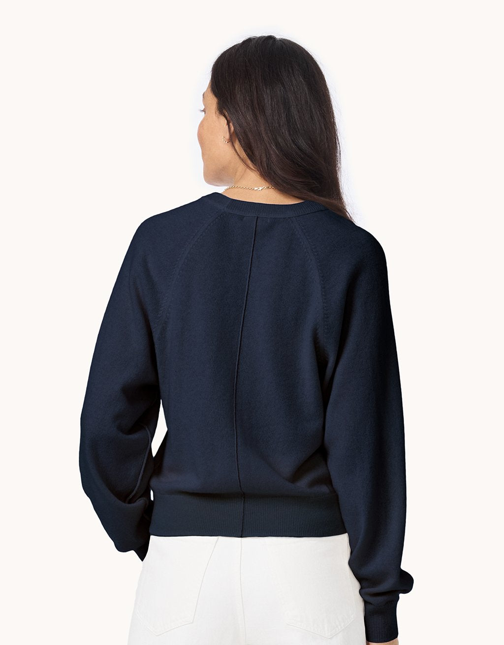 Back view of a woman wearing a navy cashmere raglan sweater and white jeans on a light gray background