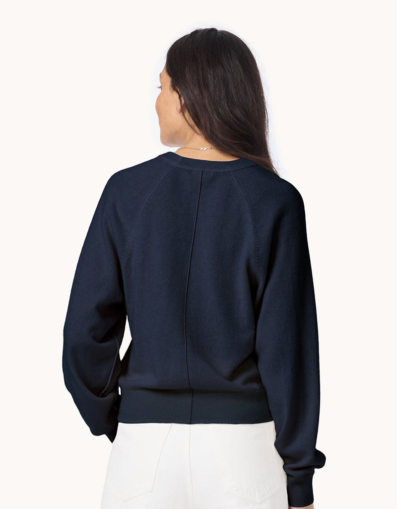 Back view of a woman wearing a navy cashmere raglan sweater and white jeans on a light gray background