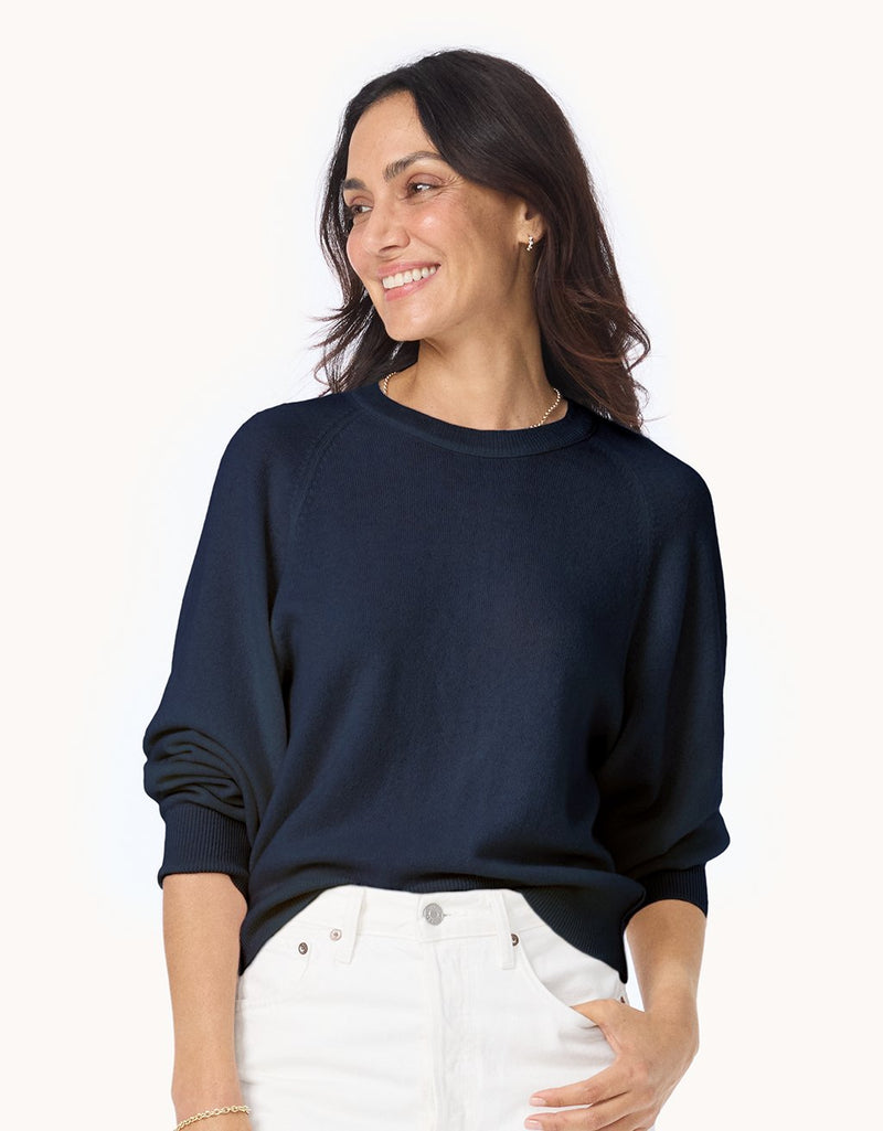 Woman wearing a navy cashmere raglan sweater and white jeans on a light gray background