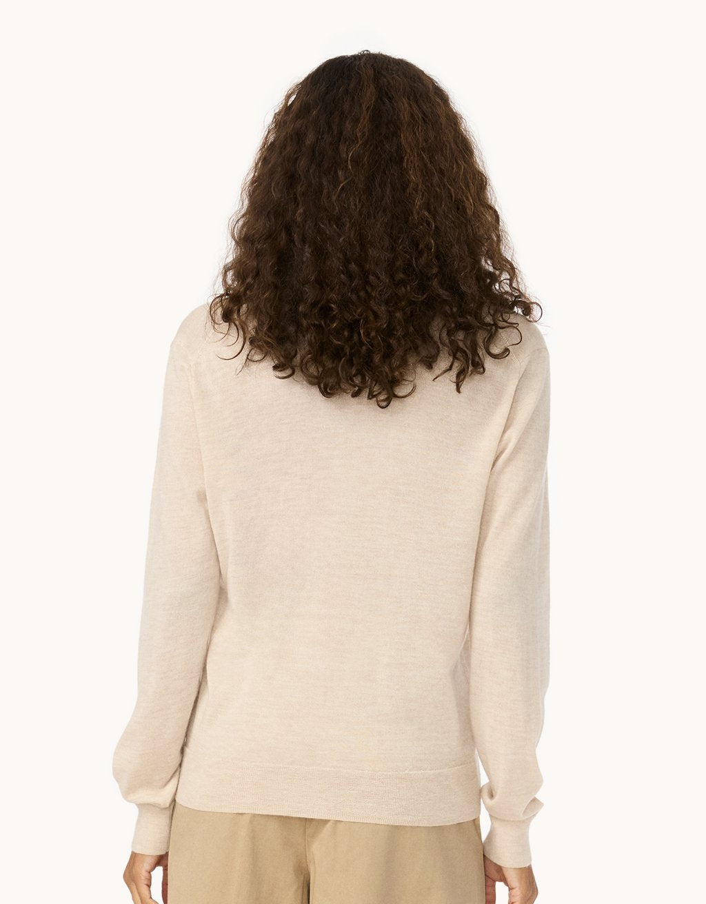Back view of a woman wearing a beige cashmere sweater and pants on a light gray background