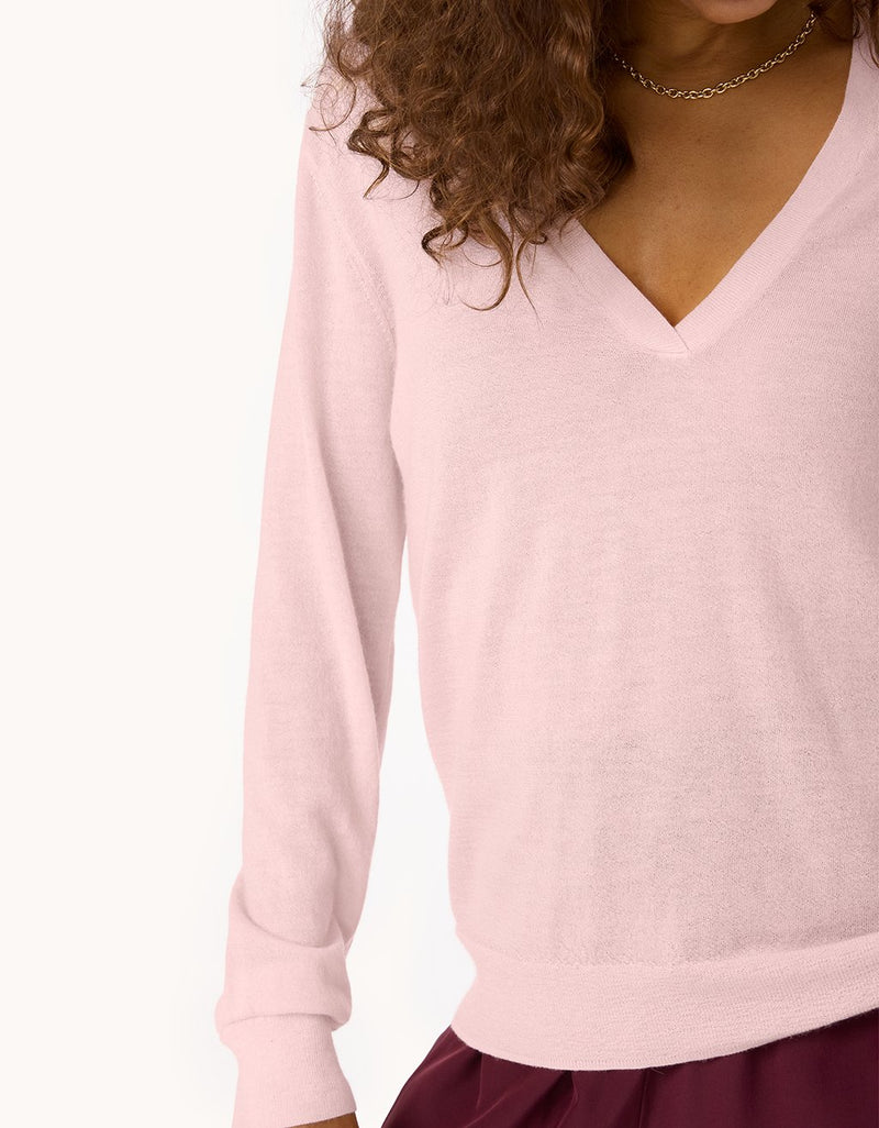 Close-up of a gold necklace, light pink cashmere V-neck sweater and maroon pants worn by a person on a light background