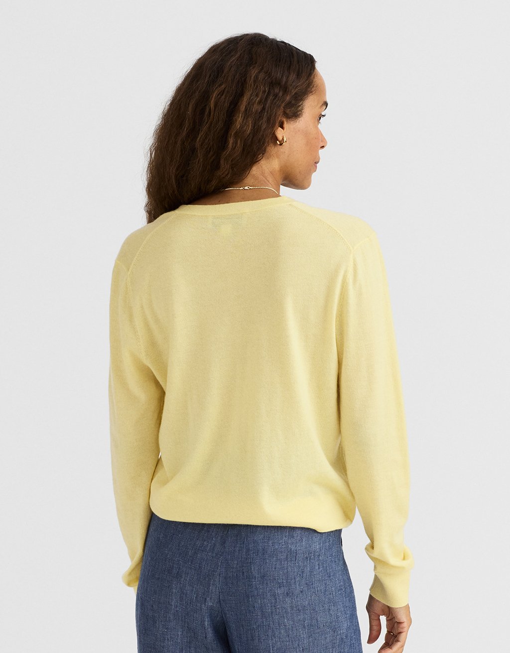 Back view of a woman wearing a light yellow cashmere sweater and blue pants on a plain background