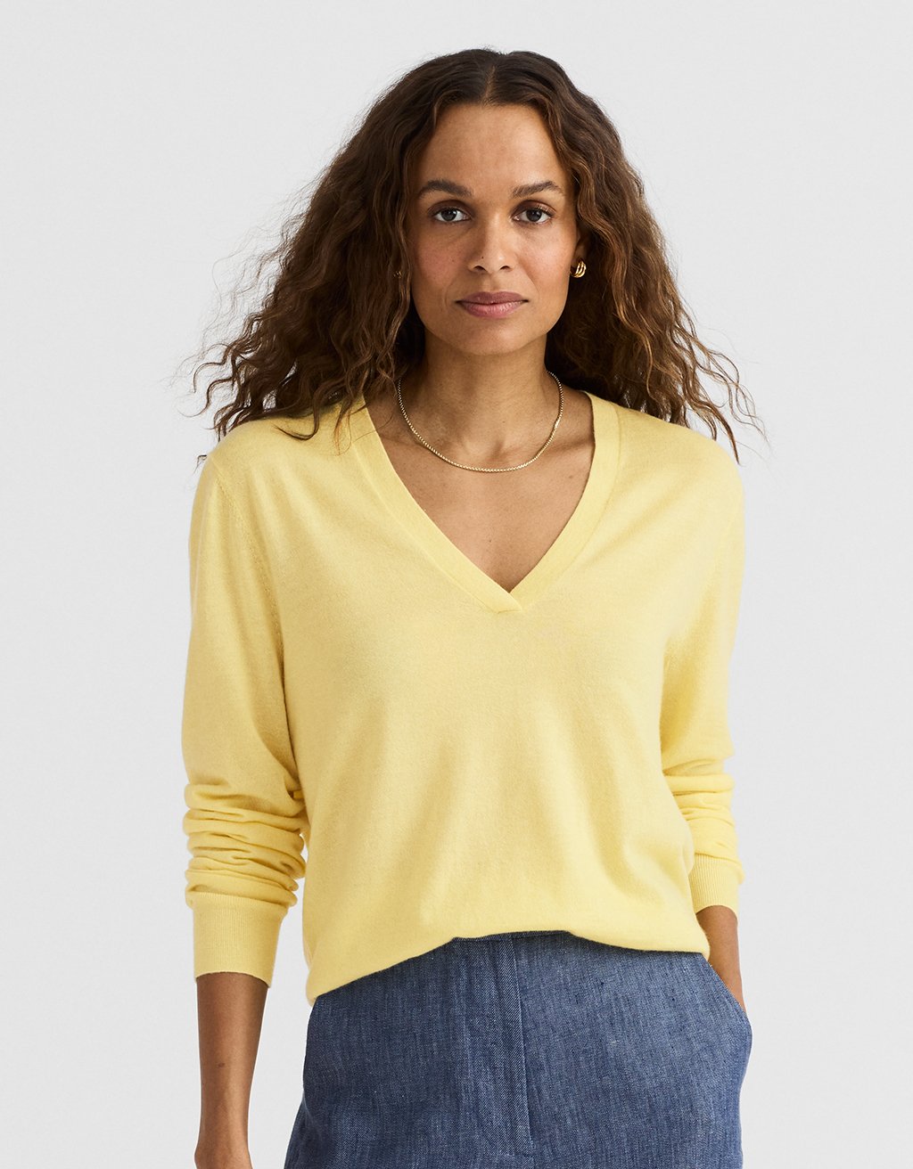 Woman wearing a yellow cashmere V-neck sweater and blue pants against a light gray background