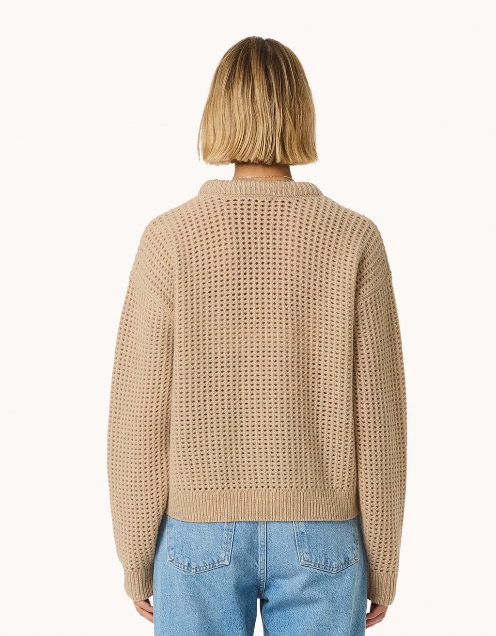 Back view of a woman wearing a camel open-stitch sweater and blue jeans on a light gray background