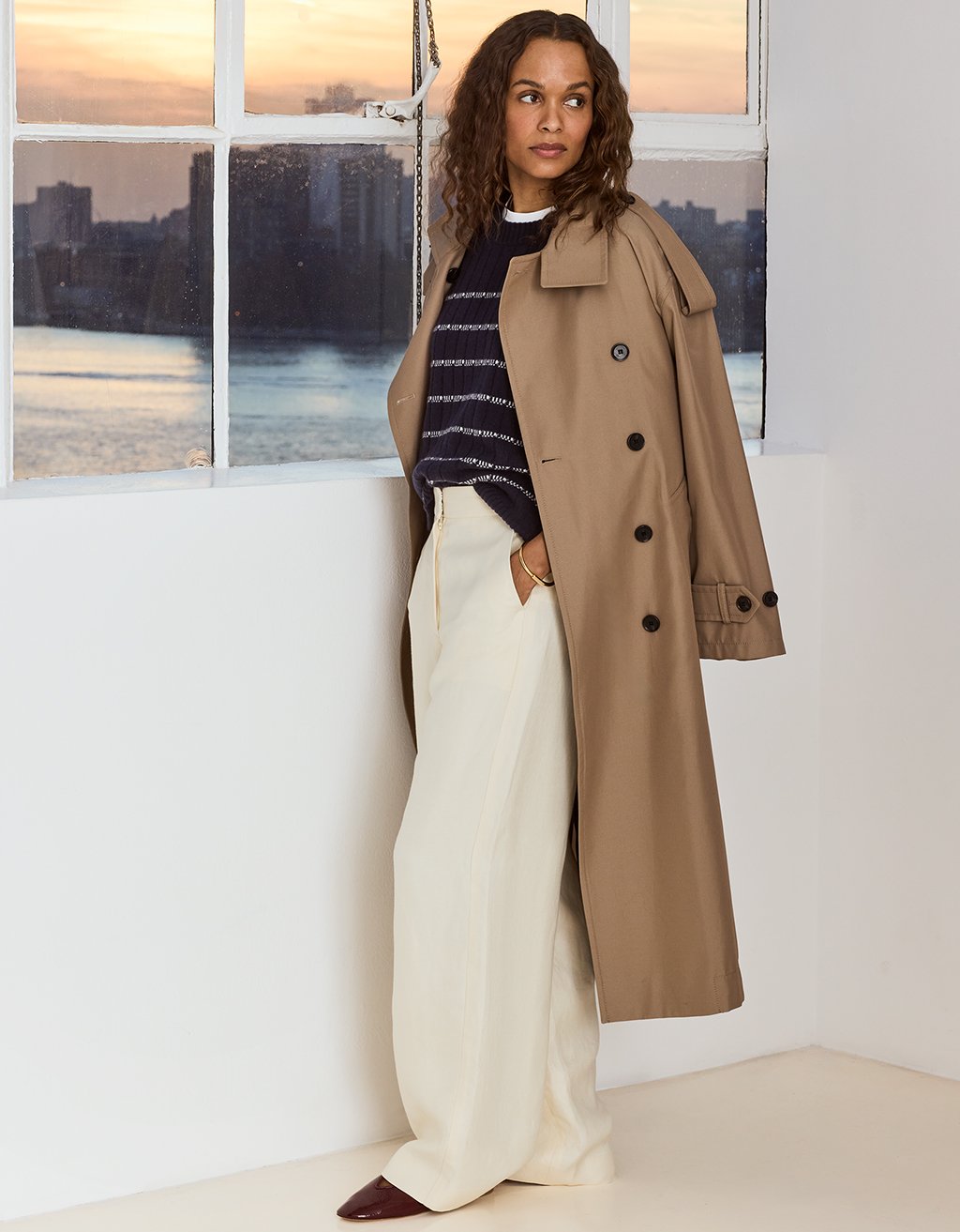 Woman wearing a beige trench coat over a navy cashmere ribbed stripe sweater with cream pants in a room with large windows showing a cityscape.
