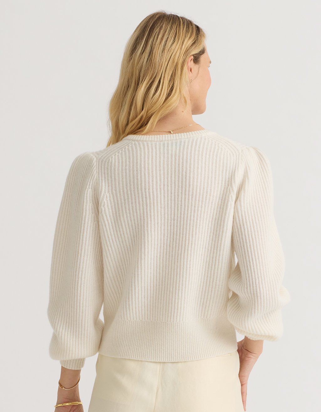 Back view of a woman wearing an ivory  cashmere puff sleeve cardigan on a light gray background