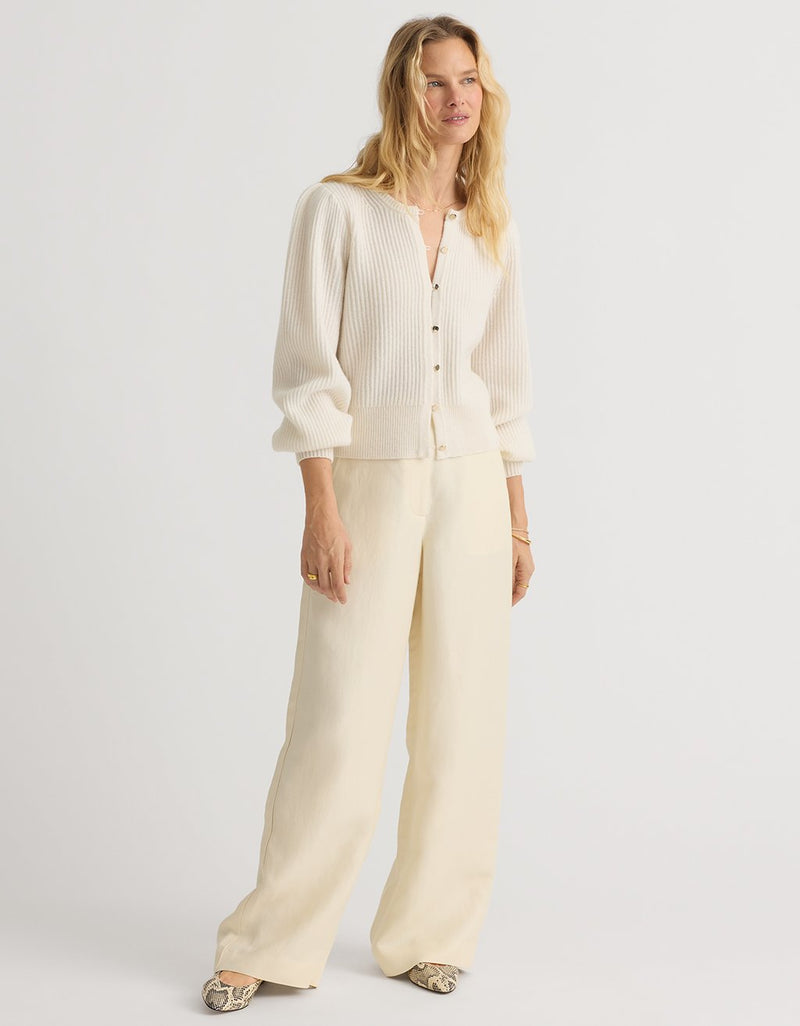 Woman wearing an ivory cashmere puff sleeve cardigan with cream pants on a light gray background