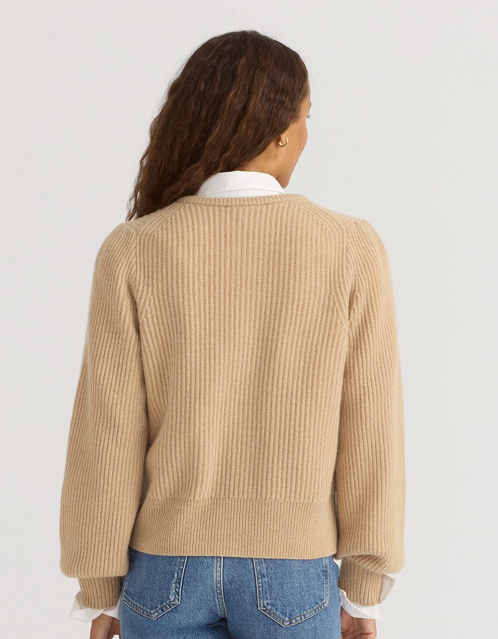 Back view of a woman wearing a camel cashmere puff sleeve cardigan and blue jeans on a plain background