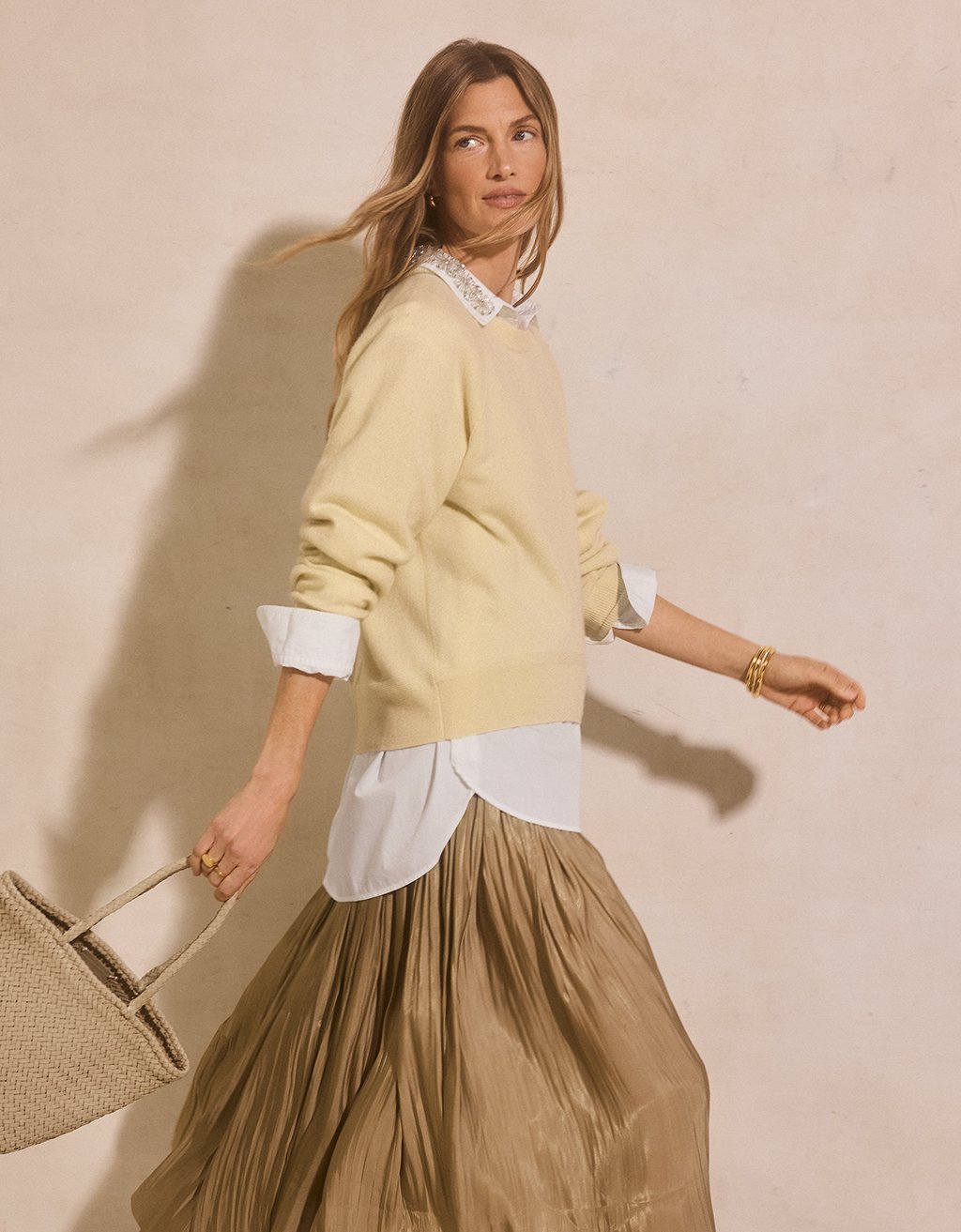 Woman wearing a light yellow cashmere raglan sweater over a white shirt with a beige skirt, holding a beige bag against a beige background.