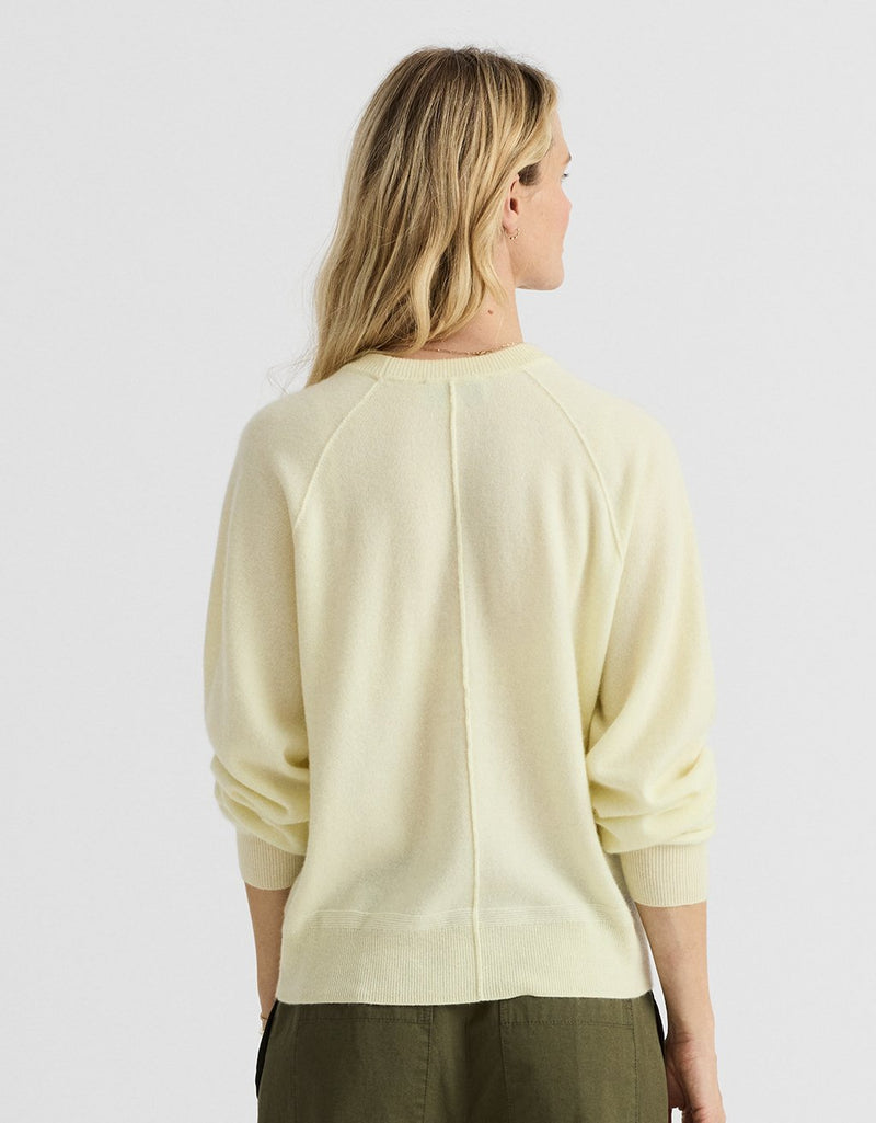 Back view of a woman wearing a light yellow cashmere raglan sweater on a light gray background