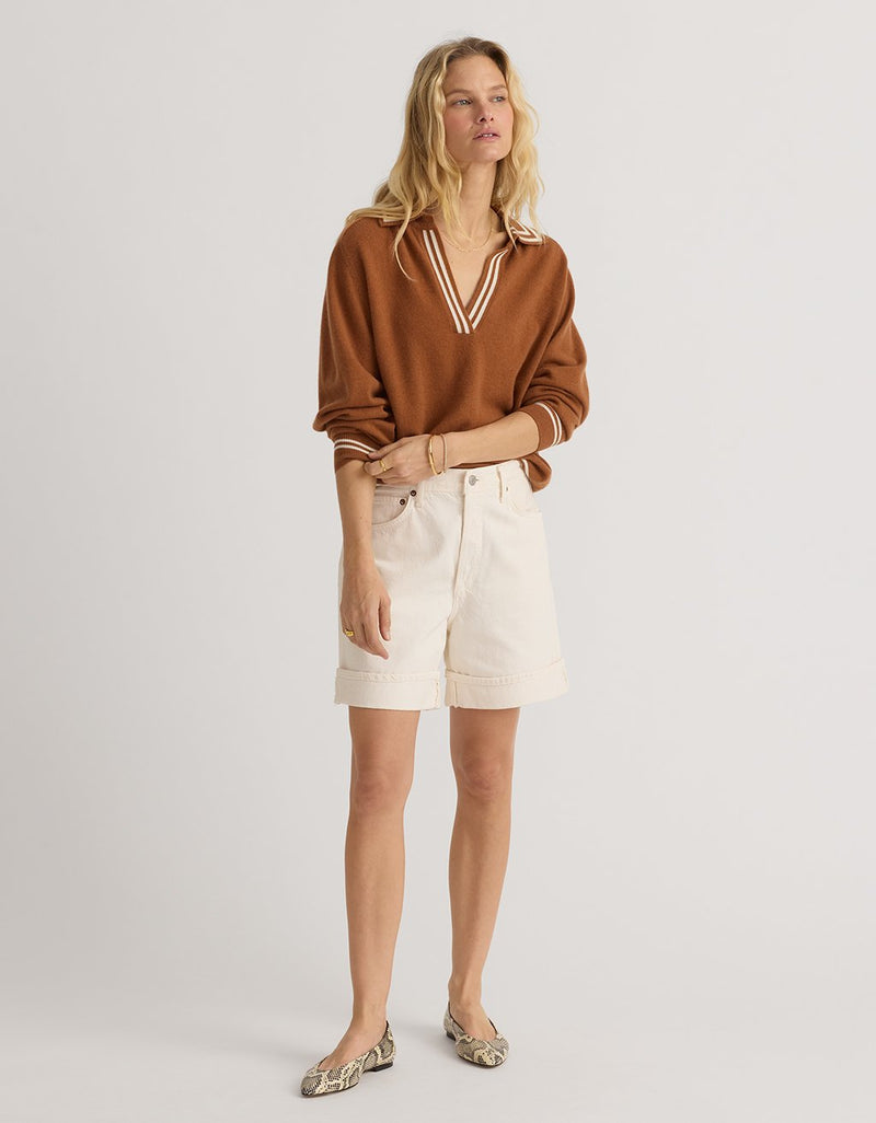 Woman wearing a burnt orange cashmere polo sweater and cream denim shorts on a plain background