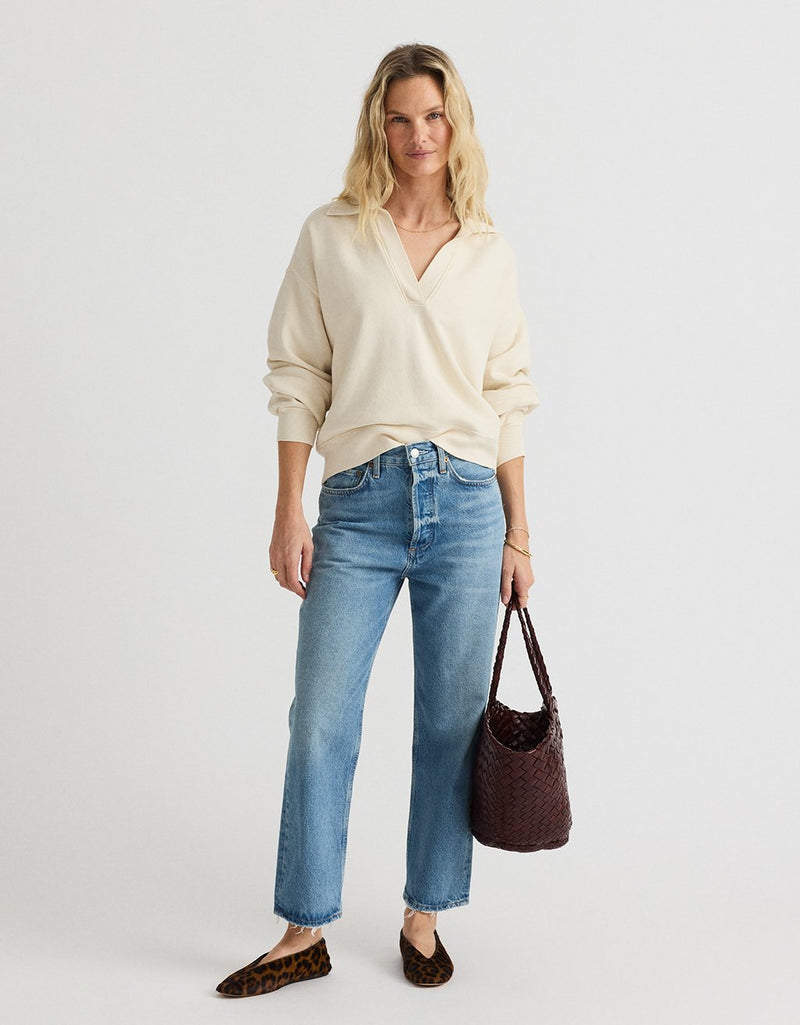 Woman wearing a cream polo sweatshirt and blue jeans, holding a woven brown bag with leopard flats