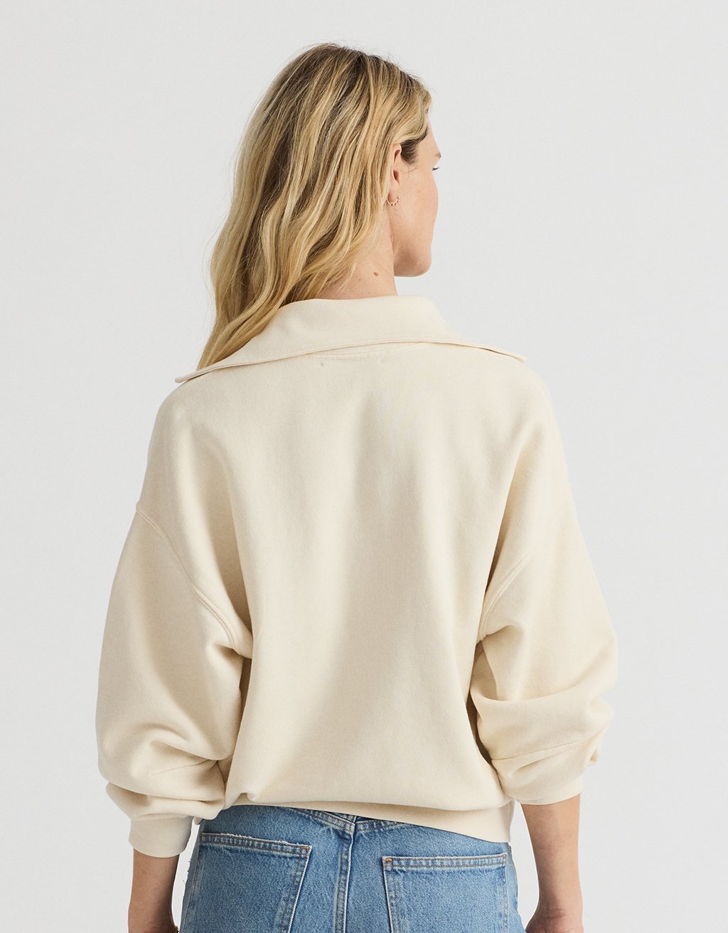 Woman wearing a cream polo sweatshirt and blue jeans on a plain background
