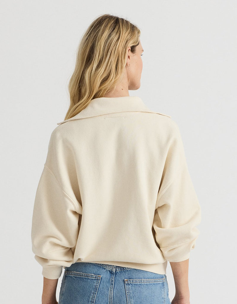 Woman wearing a cream polo sweatshirt and blue jeans on a plain background