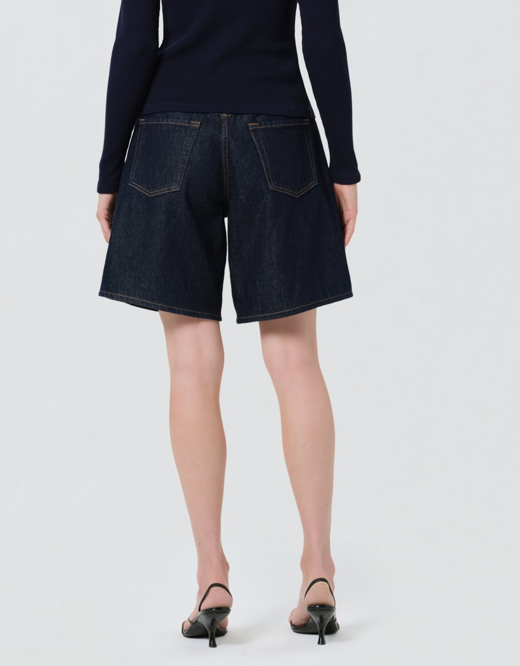 Back view of a person wearing dark denim shorts and a navy cardigan on a light gray background