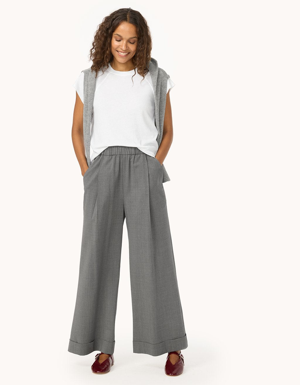 Woman wearing a white t-shirt with a gray sweater over her shoulders and wide-leg gray pants on a light gray background
