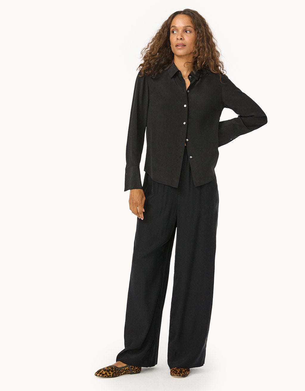 Woman wearing a black button down shirt and wide leg silk pants on a light gray background