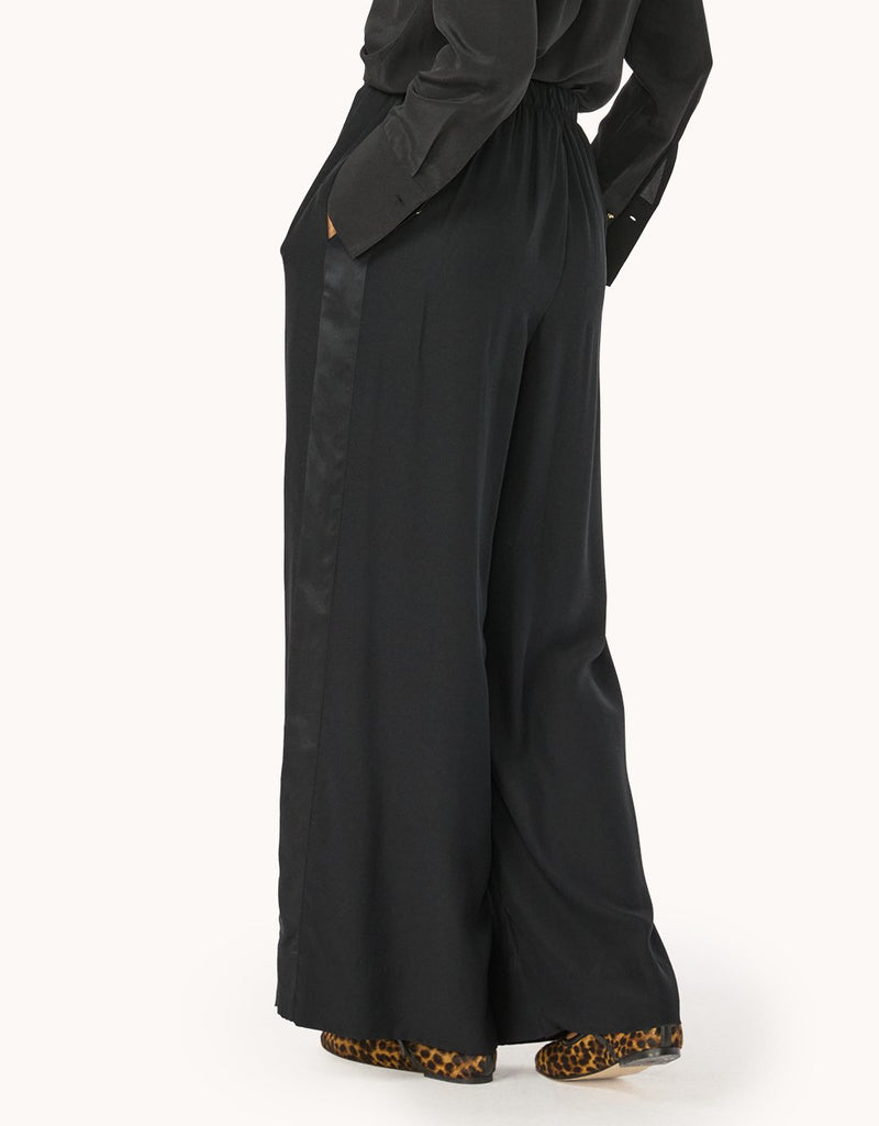 Side view of a person wearing a black top and silk wide-leg pants with a tonal side stripe detail on a light gray background