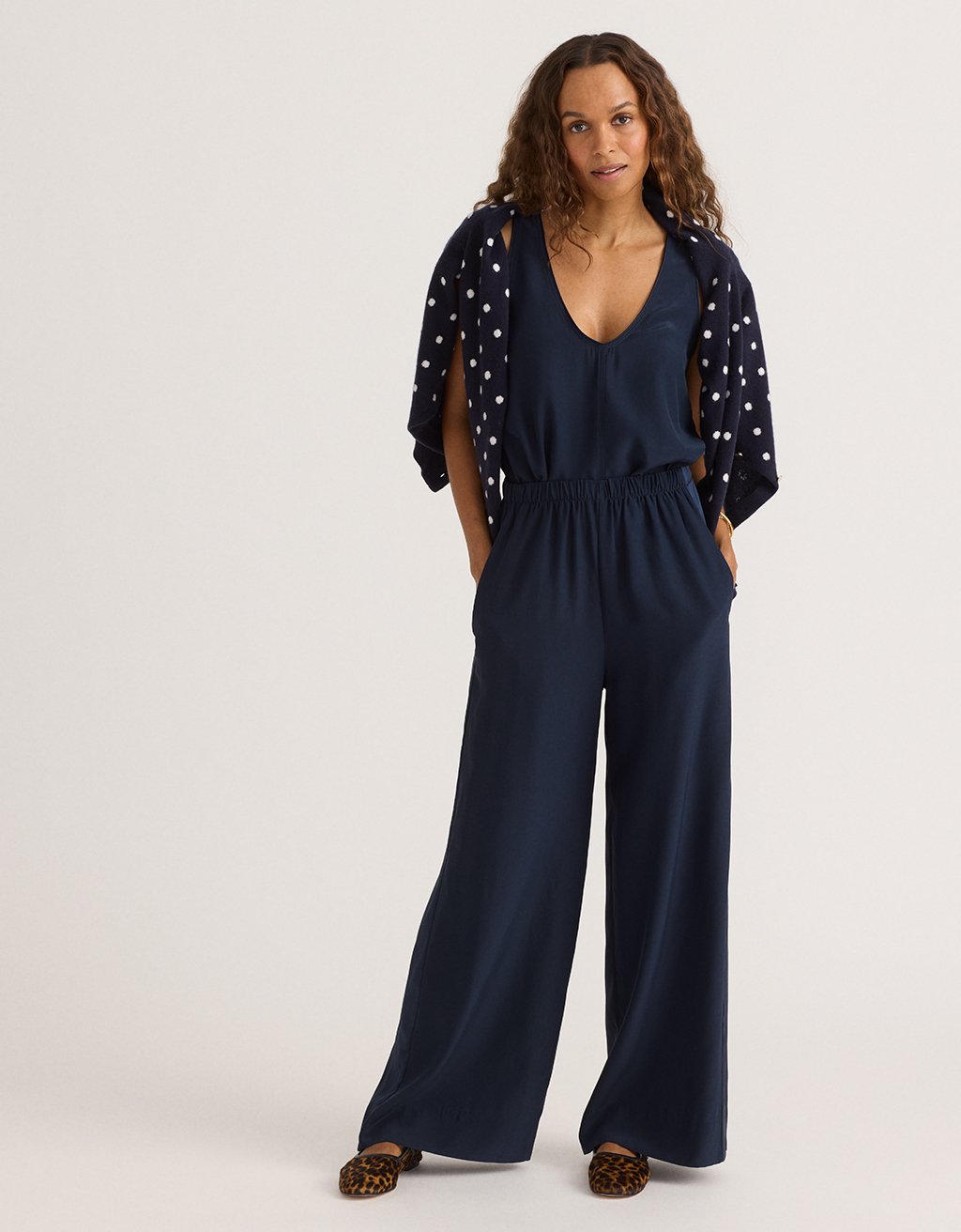 Woman wearing a navy tank and navy wide leg pants with a draped polka dot sweater on a white background