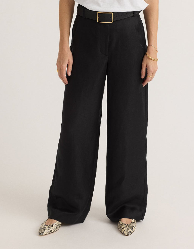 Woman wearing black wide-leg pants with a white t-shirt, gold buckle belt and snake print shoes on a light background