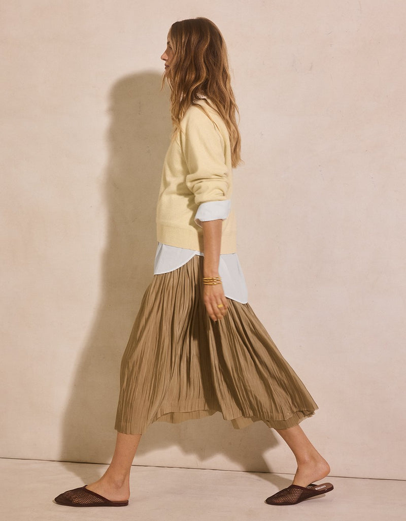 Woman wearing a light yellow cashmere sweater over a white button down shirt with a pleated olive skirt against a beige wall.