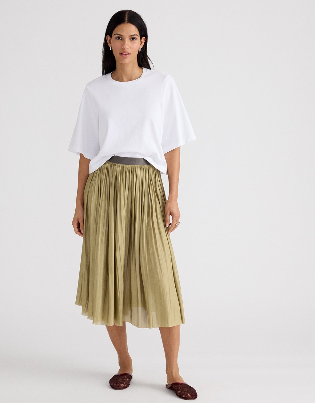 Woman sitting on a bench wearing a light yellow cashmere sweater, white shirt with jeweled collar, and olive pleated maxi skirt sitting on a bench against a beige wall
