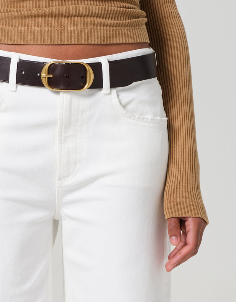 Close-up of a person wearing a brown ribbed sweater and white jeans with a  belt on a plain background.