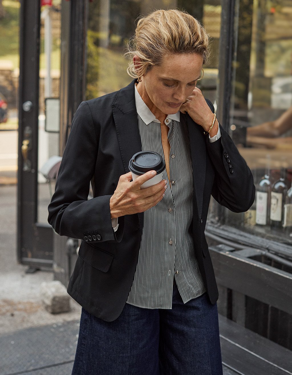 Woman in a black blazer, striped button down shirt and wide leg jeans holding a coffee cup on a city street.