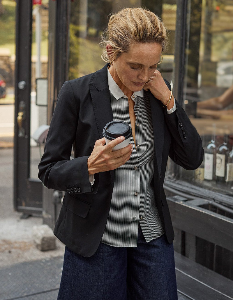 Woman in a black blazer, striped button down shirt and wide leg jeans holding a coffee cup on a city street.