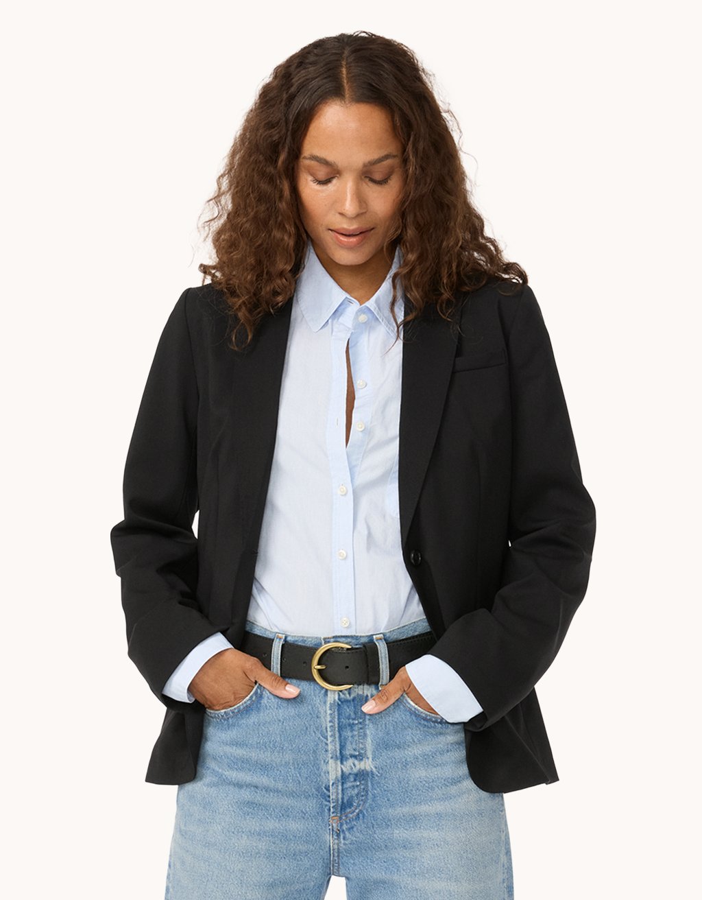 Woman wearing a black blazer, light blue button down shirt, and jeans on a light gray background