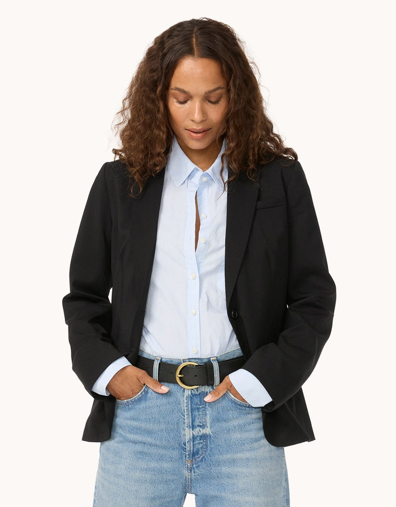 Woman wearing a black blazer, light blue button down shirt, and jeans on a light gray background
