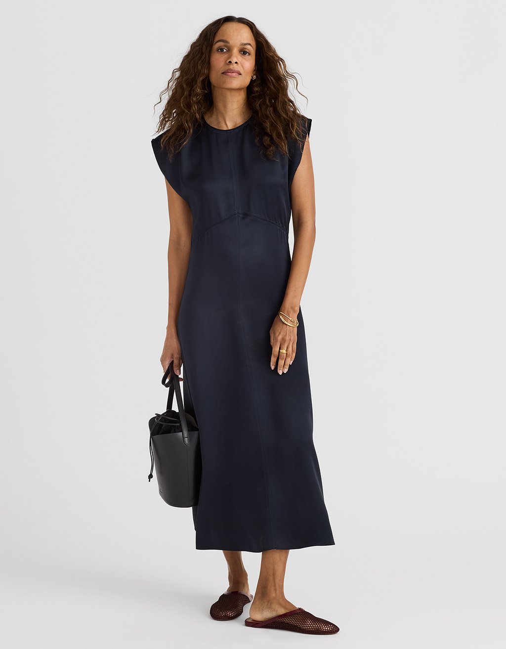 Woman wearing a navy satin midi dress holding a black handbag on a white background
