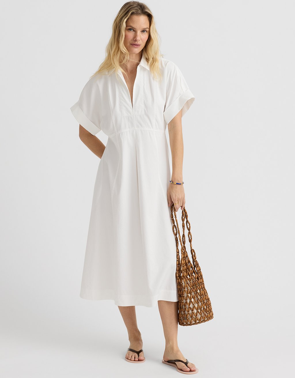 Woman wearing a white sateen pleated shirtdress holding a woven bag on a plain background