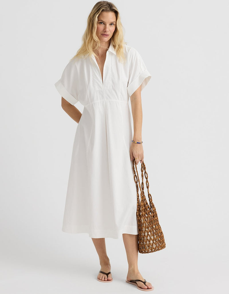 Woman wearing a white sateen pleated shirtdress holding a woven bag on a plain background